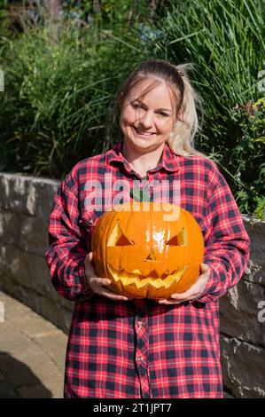 Die blonde Frau im Garten präsentiert zu Halloween einen selbst geschnitzten Kürbis mit Gesicht Stockfoto