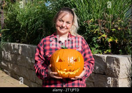 Die blonde Frau im Garten präsentiert zu Halloween einen selbst geschnitzten Kürbis mit Gesicht Stockfoto