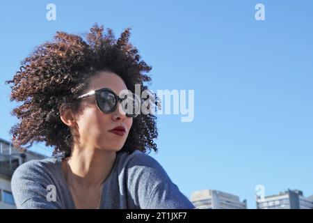 KOPFPORTRÄT EINER FRAU MIT LOCKIGEM HAAR UND SONNENBRILLE AN EINEM HEISSEN SONNIGEN TAG Stockfoto