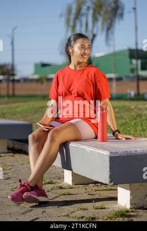 Lateinisches Mädchen in Sportbekleidung, das auf einer Bank in einem öffentlichen Park sitzt. Stockfoto