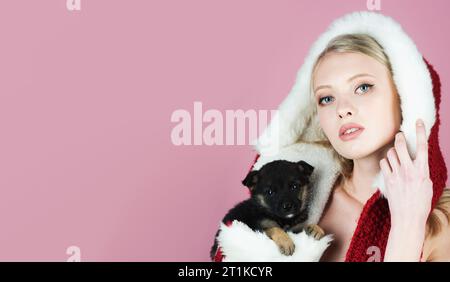 Frohe Weihnachten und frohes neues Jahr. Wunderschönes Weihnachtsmann-Mädchen mit einem kleinen Welpen. Weihnachtsfrau in Pelzmütze mit kleinem Hund. Eine junge Frau mit rotem Weihnachtsmann-Hut Stockfoto