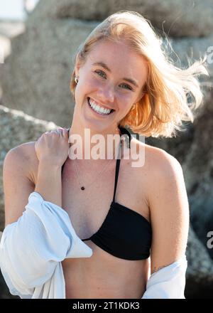 Eine junge Frau, die am Strand steht und lächelt, in einem schwarzen zweiteiligen Badeanzug Stockfoto