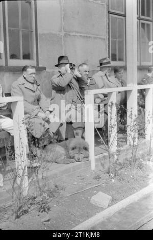 Cricket Match-Abdeckung von einer Übereinstimmung zwischen Kenton und Alexandra Park, Kenton, Middlesex, England, UK, 1945 Zuschauer, darunter ein Hund, ein kleiner Junge und ein Mitglied der RAF, sitzen auf Bänken vor dem Pavillon im Sonnenschein ein Cricket-spiel zwischen Kenton und Alexandra Park an der Kenton Boden, Harrow, Middlesex zu beobachten. Ist ein Mann mit Fernglas den Fortschritt des Spiels mehr genau zu verfolgen. Stockfoto