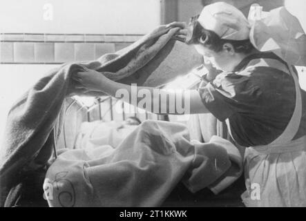 Guy's Hospital - Leben in einem Londoner Krankenhaus, England, 1941 EIN VAD (freiwillige Hilfe Loslösung) Krankenschwester ordnet eine Asbest Decke über ein elektrisch beheiztes Frame eine Kapuze über diesen Patienten zu erstellen, um Ihnen zu helfen, schnell warm. In den Fällen der schweren Schock, Kleidung des Patienten sind abgeschnitten, so dass die Oberfläche der Haut können einige in engeren Kontakt mit der Hitze. Stockfoto