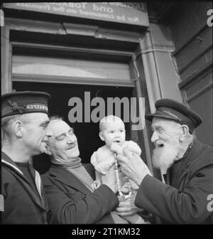 Heilige Insel spielt seine Rolle - Alltag auf Lindisfarne, 1942 George Allison (Zweiter von rechts), der Vermieter der Krone und Anker Public House auf Lindisfarne, zeigt ein Baby (vielleicht sein Enkel) in ein alter Fischer und Mitglied einer Minesweeper Crew, außerhalb des Inn. Die Fischer hilft, das Baby mit einer Zeichenkette der Große Perlen zu spielen. George ist 64 Jahre alt und ist der Sergeant der Fischer Home Guard. Stockfoto