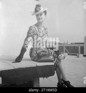 Wie eine britische Frau Kleider in Kriegszeiten - Utility Kleidung in Großbritannien, 1943 ein Modell, das sich auf eine niedrige Mauer auf einem Bloomsbury Dachterrasse sitzt in ihr vor dem Krieg gedruckt Seidenkleid zeigen. Gemäß der ursprünglichen Bildunterschrift, dieses Kleid wurde "olled" durch, mit schwarzen Handschuhen und Handtasche getragen und auch mit einem weißen Hut mit roten Geranien und blauen Kornblumen, die Farbe des Drucks der Kleid zu passen. Stockfoto