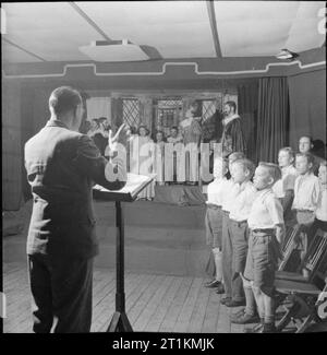 Lernen durch Handeln - Bildung durch Theater für Umsiedler in einem 'Camp Schule', 'sMarchant Hill, Hindhead, Surrey, 1943 Musik Lehrer Arthur Shuttleworth (zurück in die Kamera) Führt der Chor (rechts) und Cast (auf der Bühne im Hintergrund) in einer Auswahl von traditionellen englischen Songs, das Stück über elisabethanischen England, die Sie in der pausenhalle an der Marchant Hill School, Hindhead gesetzt haben, zu schließen. Die Kinder haben von East Ham evakuiert und alle haben um die Leistung in irgendeiner Weise beigetragen. Deutlich sichtbar auf der Bühne, als Königin Elizabeth I, ist der 13 Jahre alte Mavis Clode. Stockfoto