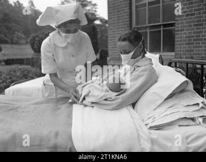 Herrenhaus wird Mutterschaft Startseite - Leben bei Brocket Hall, Welwyn, Hertfordshire, 1942 eine Krankenschwester Prüfungen auf einer Mutter als sie wiegt Ihr Baby auf ein Bett in der Luft außerhalb Brocket Hall. Beide Frauen sind das Tragen von Atemschutzmasken die Ausbreitung von Keimen auf den Säugling zu vermeiden. Stockfoto