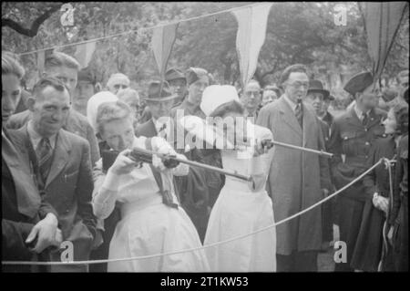 Krieg Fair - Urlaub zu Hause auf einer Fete in Russell Square, London, 1943 Zwei Krankenschwestern gehen an ein Ballerspiel haben auf einer Messe in Russell Square. Ziel ist es, die Zigarettenpackungen mit Korken zu schießen. Eine große Gruppe von Menschen, einschließlich der RAF und Armeeangehörigen, Uhren, wie sie Feuer. Bunting schmückt den Platz hinter Ihnen. Stockfoto