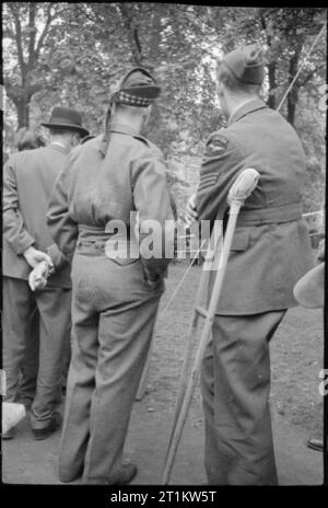 Krieg Fair - Urlaub zu Hause auf einer Fete in Russell Square, London, 1943 eine Gruppe von drei Männern, einer zivilen, eine von einem schottischen Regiments und ein Drittel von der Royal Air Force, stand mit dem Rücken zur Kamera, wie sie die Festlichkeiten stattfinden am Russell Square fete beobachten. Die RAF-Mann auf Krücken. Stockfoto