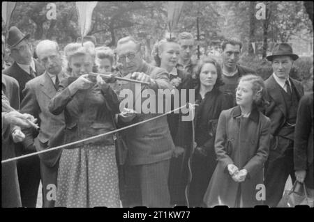 Krieg Fair - Urlaub zu Hause auf einer Fete in Russell Square, London, 1943 eine Frau spielt ein Spiel auf der Messe in Russell Square. Sie ist von einem Mann, der erklärt, dass Sie die Korken, mit denen die Waffe auf Zigaretten geladen ist Schießen unterstützt. Andere Mitglieder der Öffentlichkeit. Bunting können nur im Hintergrund gesehen werden. Stockfoto