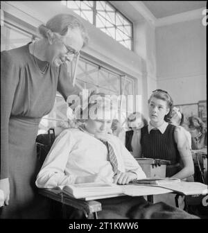 Young Britons studieren amerikanische Geschichte - Ausbildung in Kriegszeit England, 1943 Geschichtslehrerin Miss L M Molineux hilft Monica Young bei ihrer Arbeit während einer Lektion über amerikanische Geschichte an der Albany Senior School in Enfield. Monica ist behindert und hat einen Schreibtisch auf den Armen ihres Rollstuhls, auf dem sie schreiben kann. Die Glasscheiben, die den Klassenzimmer vom Flur trennen, sind mit Klebeband versehen, um Schäden durch Explosionen zu verhindern. Stockfoto