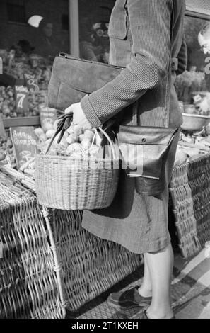 Eine Hausfrau mit Warenkorb und Gasmaske Behälter Geschäfte zu einem Obst- und Gemüsehändler in London 1941. Eine Nahaufnahme der Warenkorb von Frau Tag und Gasmaske bei, die Sie auf Ihrem Einkaufsbummel getroffen hat. Die Gasmaske Fall hat eine spezielle Tasche, der es ihr erlaubt, wie eine Handtasche verwendet werden. Hinter ihr, der gemüsehändler ordnet seine Waren. Dieses Foto wurde wahrscheinlich auf der King's Road in Chelsea. Stockfoto