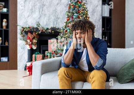 Starke Kater Kopfschmerzen und Migräne, Mann mit Kopf sitzend auf Sofa im Wohnzimmer zu Weihnachten, afroamerikanischer Mann mit lockigen Haaren in der Nähe von dekoriertem Weihnachtsbaum. Stockfoto