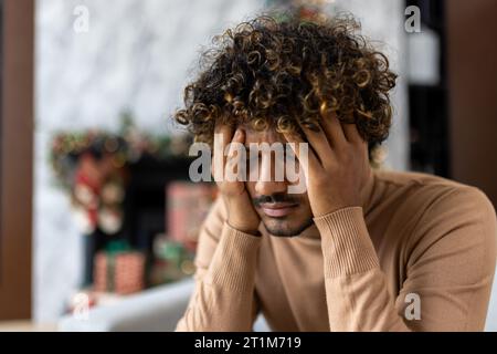 Starke Kater Kopfschmerzen und Migräne, Mann mit Kopf sitzend auf Sofa im Wohnzimmer zu Weihnachten, afroamerikanischer Mann mit lockigen Haaren in der Nähe von dekoriertem Weihnachtsbaum. Stockfoto