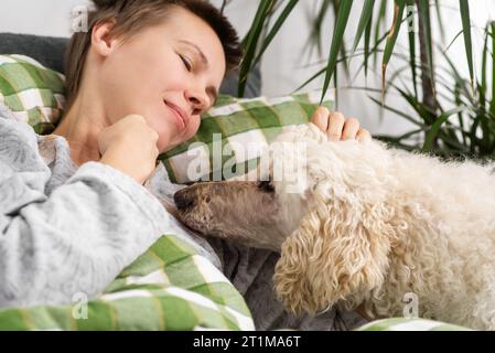 Den ganzen Tag hindurch liegt eine Frau mittleren Alters mit kurzen Haaren im Bett mit ihrem großen Pudel. Inaktivität, entspanntes Wochenende Stockfoto