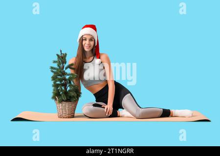 Sportliche junge Frau im Weihnachtsmann-Hut mit Weihnachtsbaum auf blauem Hintergrund Stockfoto