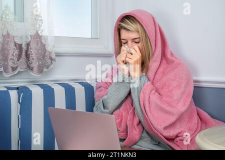 Ein Mädchen niest in einer Decke und benutzt zu Hause einen Laptop Stockfoto
