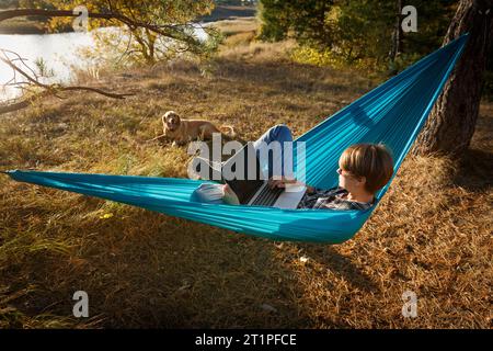 Junge Frau im Hammok, die mit ihrem Laptop im Freien arbeitet und mit Blick auf den See bei Sonnenuntergang zusammen mit ihrem Haustierhund arbeitet. Konzept der Remote-Jobchance Stockfoto