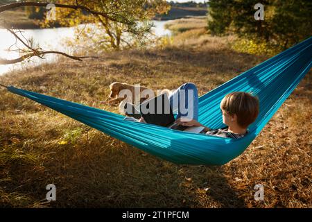 Junge Frau im Hammok, die mit ihrem Laptop im Freien arbeitet und mit Blick auf den See bei Sonnenuntergang zusammen mit ihrem Haustierhund arbeitet. Konzept der Remote-Jobchance Stockfoto