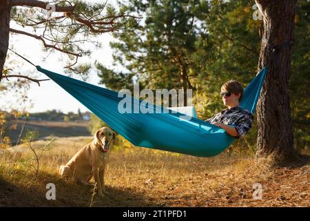 Junge Frau im Hammok, die mit ihrem Laptop im Freien arbeitet und mit Blick auf den See bei Sonnenuntergang zusammen mit ihrem Haustierhund arbeitet. Konzept der Remote-Jobchance Stockfoto