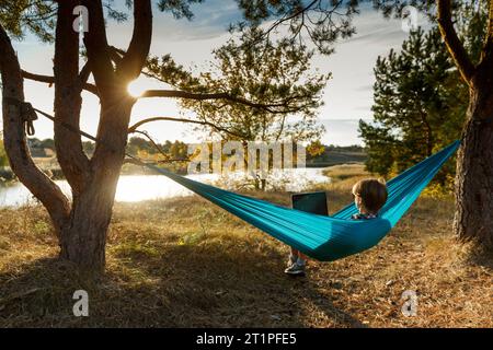 Junge Frau im Hammok mit Laptop im Freien mit Blick auf den See bei Sonnenuntergang. Konzept der Remote-Jobchance Stockfoto