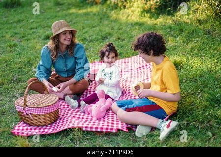 Die fröhliche Mutter und ihre beiden Kinder genießen einen sonnigen Tag mit einem Picknick auf dem grünen Gras, umgeben von der Natur. Stockfoto