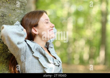 Profil einer glücklichen Frau, die sich im Wald auf einen Baum lehnt Stockfoto