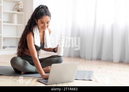 Glückliche indianerin, die eine Pause vom virtuellen Workout macht und Wasser trinkt Stockfoto