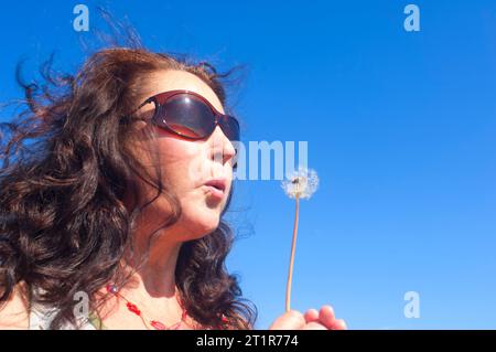 Reife Frau, die eine Löwenzahnuhr bläst - John Gollop Stockfoto