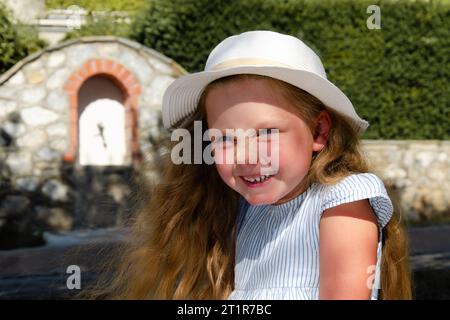 Ein Mädchen von etwa fünf Jahren in einer weißen Mütze mit lockeren blonden Haaren lacht überrascht. Porträt von der Taille nach oben. Weißes Mauerwerk und Grün in t Stockfoto