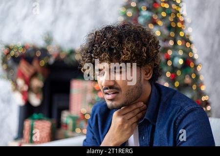 Ein Mann hat zu Weihnachten Halsschmerzen, hispanic sitzt allein zu Hause im Wohnzimmer, hält seine Kehle mit Tränen, hat eine Erkältung, hat starke Schmerzen, einen geschmückten Weihnachtsbaum mit Geschenken. Stockfoto