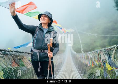 Junge lächelnde Frau überquerte den Canyon über die Hängebrücke und berührte bunte tibetische Gebetsfahnen über der Schlucht. Kletterroute auf dem Gipfel Mera Stockfoto