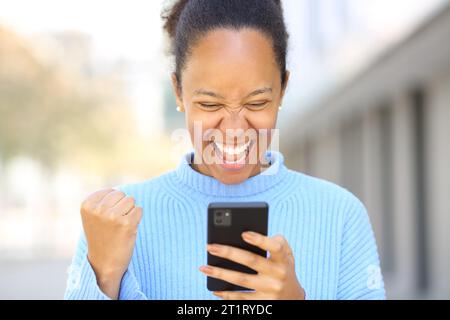 Vorderansicht Porträt einer aufgeregten schwarzen Frau, die auf der Straße gute Nachrichten am Telefon überprüft Stockfoto