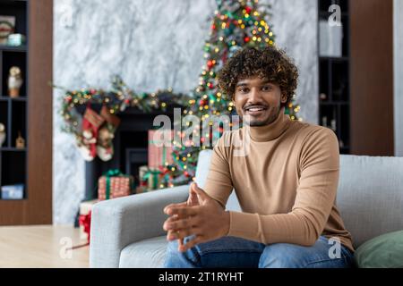 Weihnachtsportrait des jungen glücklichen Mannes, hispanischer Mann lächelnd und in die Kamera schauend, fröhlich und feiernd Weihnachten sitzend auf Sofa im Wohnzimmer in der Nähe von dekoriertem Weihnachtsbaum. Stockfoto