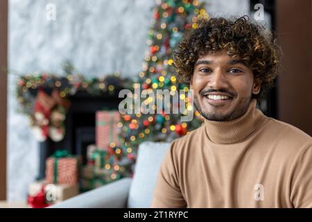 Weihnachtsportrait des jungen glücklichen Mannes, hispanischer Mann lächelnd und in die Kamera schauend, fröhlich und feiernd Weihnachten sitzend auf Sofa im Wohnzimmer in der Nähe von dekoriertem Weihnachtsbaum. Stockfoto