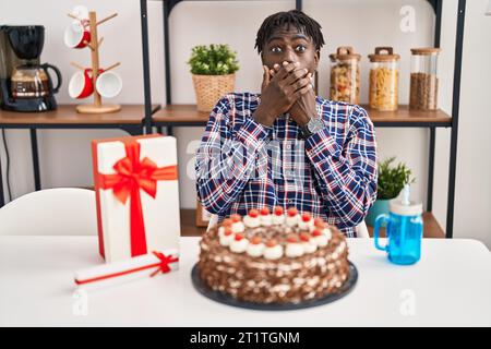 Afrikanischer Mann mit Dreadlocks, der Geburtstag feiert und großen Schokoladenkuchen schockiert hält, der den Mund aus Versehen mit Händen bedeckt. Geheimes Konzept. Stockfoto
