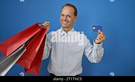 Ein selbstbewusster, lächelnder Mann mittleren Alters genießt es, sich bei einem modischen Kauf hervorzuheben, indem er Einkaufstaschen und Kreditkarten hält und isoliert positioniert wird Stockfoto