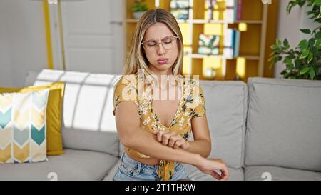 Junge blonde Frau sitzt auf dem Sofa und kratzt den Arm, weil sie zu Hause juckt Stockfoto