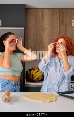 Mutter und Tochter machen lustige Gesichter mit geformten Keksformen, während sie Butterkekse zubereiten. Familienzeit. Stockfoto