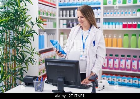 Junge schwangere Apothekerin lächelt selbstbewusst mit Computer und Touchpad in der Apotheke Stockfoto