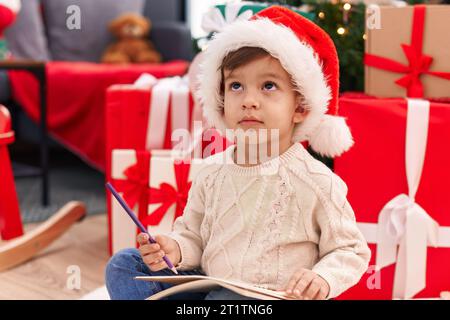 Ein entzückender hispanischer Junge, der den weihnachtsmann-Brief schreibt, sitzt zu Hause am weihnachtsbaum auf dem Boden Stockfoto