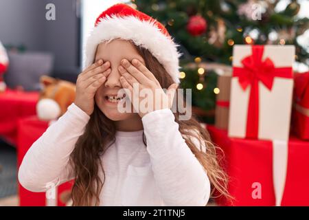 Entzückendes hispanisches Mädchen, das die Augen mit Händen bedeckt, die zu Hause am weihnachtsbaum auf dem Boden sitzen Stockfoto