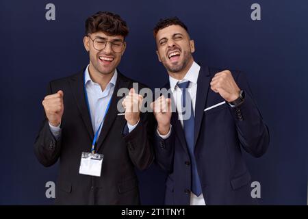 Zwei hispanische Geschäftsmänner in Geschäftskleidung, die mit erhobenen Armen und geschlossenen Augen zum Erfolg aufgeregt sind und lächelnd den Sieg feiern. Gewinnerkonzept. Stockfoto