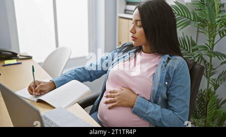 Erwartende junge hispanische Geschäftsfrau, Aufgabenjonglieren in ihrem Beruf, sensibles Gleichgewicht zwischen Schwangerschaft und Arbeit - Notizen auf ihrem Laptop im Handumdrehen Stockfoto