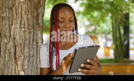 Coole afroamerikanische Frau, die sich auf ihr Touchpad fokussiert, sich im Stadtpark auf einen Baum lehnt, lässige Lifestyle-Atmosphäre im Grünen Stockfoto