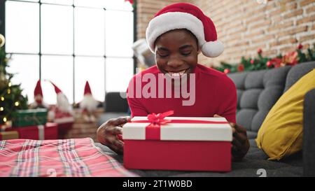 afroamerikanerin trägt weihnachtsmütze und packt ein Geschenk mit fröhlichem Gesicht zu Hause aus Stockfoto