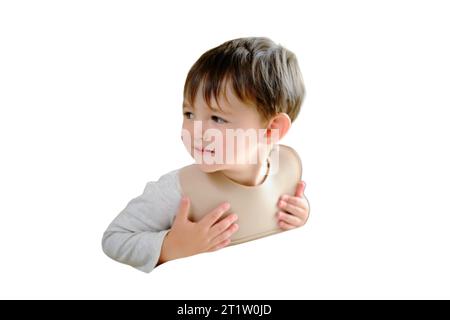 Das Kind wartet ungeduldig auf Essen, das am Tisch sitzt, in einem Lätzchen, isoliert auf weißem Hintergrund. Junge im Alter von zwei Jahren (zweijährig) Stockfoto