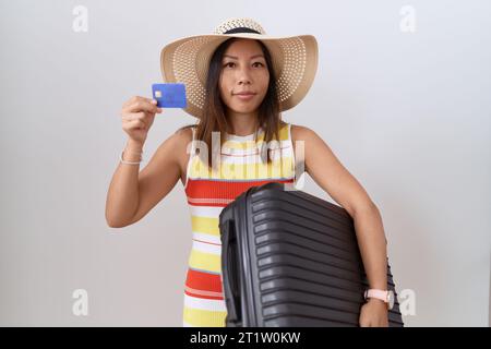 chinesische Frau mittleren Alters, die Koffer und Kreditkarte entspannt hält, mit ernstem Gesichtsausdruck. Einfaches und natürliches Betrachten der Kamera. Stockfoto