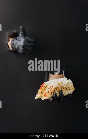 Die Hand hält den Döner und zeigt den Daumen nach oben durch gerissene Löcher auf schwarzem Papierhintergrund Stockfoto